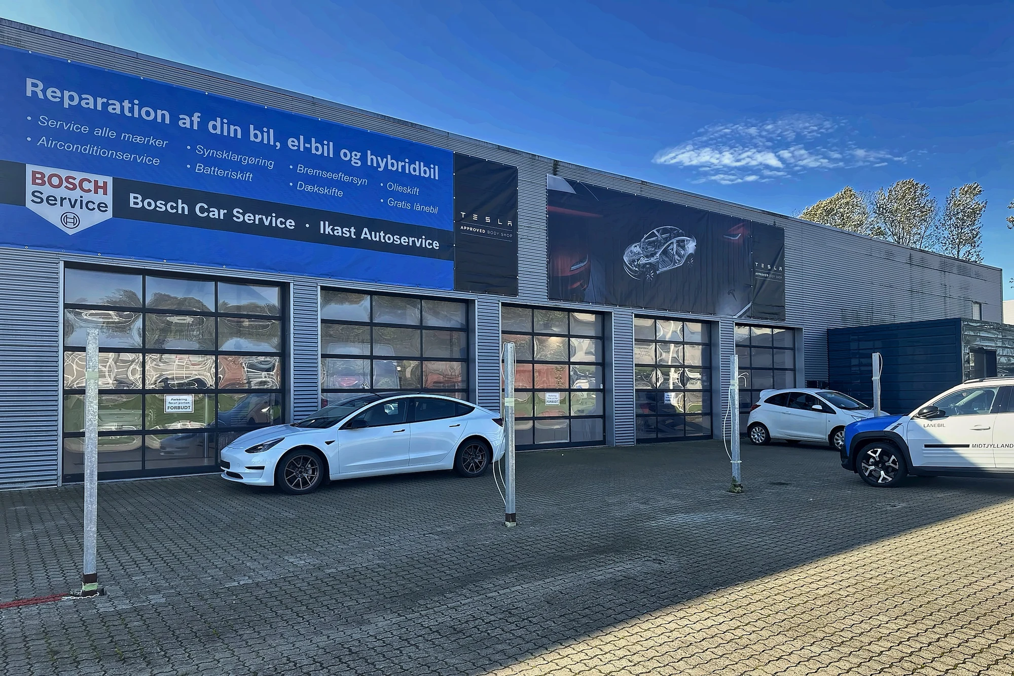 Ikast AutoService set skråt udefra, med Bosch Car Service-banner og Tesla Approved Body Shop-banner